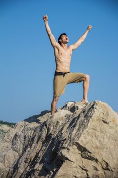 Successful sexy muscular shirtless young man with fists raised, shouting for joy, happy, standing on big rock with blue sky behind him