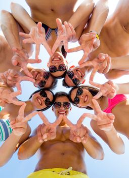 friendship, happiness, summer vacation, holidays and people concept - group of smiling friends wearing swimwear and showing victory or peace sign standing in circle over blue sky