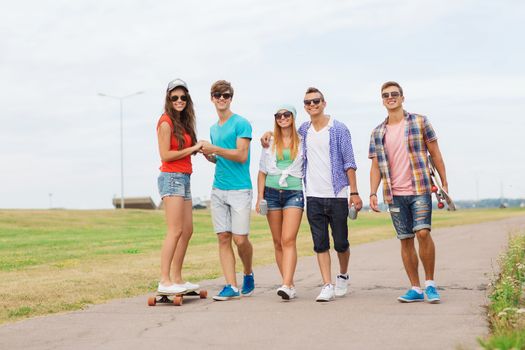 holidays, vacation, love and friendship concept - group of smiling teenagers walking and riding on skateboards outdoors