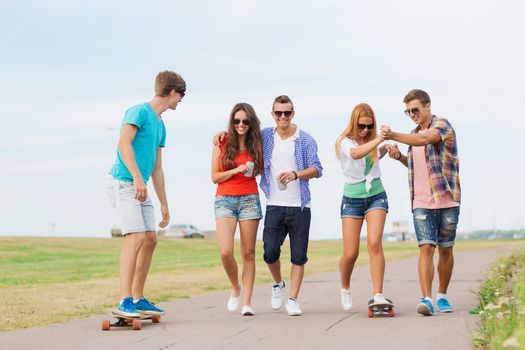 holidays, vacation, love and friendship concept - group of smiling teenagers walking and riding on skateboards outdoors