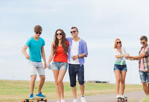 holidays, vacation, love and friendship concept - group of smiling teenagers walking and riding on skateboards outdoors