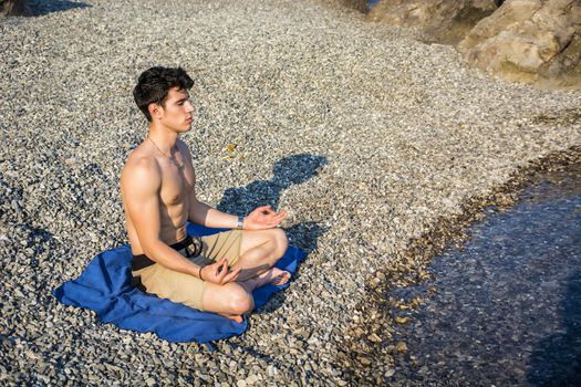 Handsome Shirtless Young Man During Meditation or Doing an Outdoor Yoga Exercise Sitting on Rocks by Ocean Shore