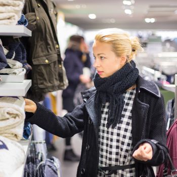 Woman shopping clothes. Shopper looking at clothing indoors in store. Beautiful blonde caucasian female model wearing casual winter clothing and black scarf.