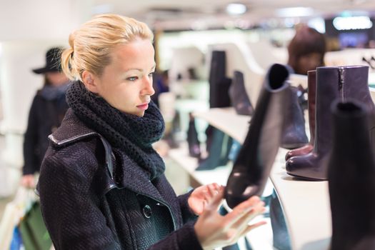 Woman shopping shoes. Shopper looking at ancle high boots on display shelf in shoe store. Beautiful blonde caucasian female model. 