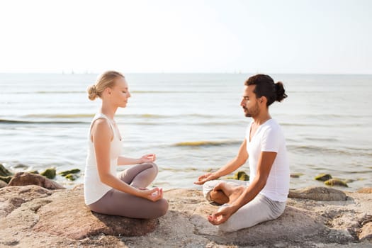 fitness, sport, friendship and lifestyle concept - smiling couple making yoga exercises sitting on sand outdoors