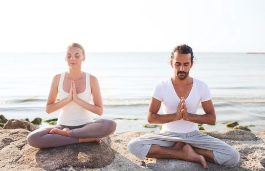 fitness, sport, friendship and lifestyle concept - smiling couple making yoga exercises sitting on sand outdoors