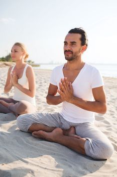 fitness, sport, friendship and lifestyle concept - smiling couple making yoga exercises sitting on sand outdoors