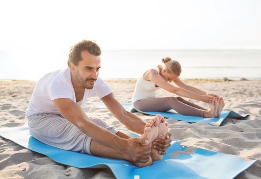 fitness, sport, friendship and lifestyle concept - couple making yoga exercises sitting on mats outdoors