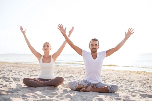 fitness, sport, friendship and lifestyle concept - smiling couple making yoga exercises sitting on sand outdoors