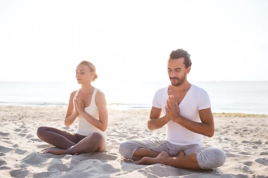 fitness, sport, friendship and lifestyle concept - smiling couple making yoga exercises sitting on sand outdoors