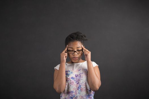 South African or African American black woman teacher or student holding her fingers on her temples thinking against a chalk black board background inside