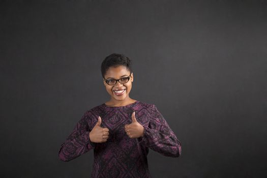 South African or African American black woman teacher or student with a thumbs up hand signal standing against a chalk blackboard background inside