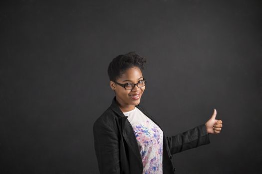 South African or African American black woman teacher or student with a thumbs up hand signal standing against a chalk blackboard background inside