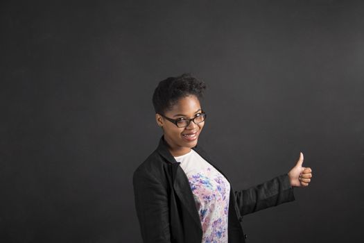 South African or African American black woman teacher or student with a thumbs up hand signal standing against a chalk blackboard background inside