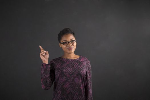 South African or African American black woman teacher or student with a good idea or answer standing against a chalk blackboard background inside