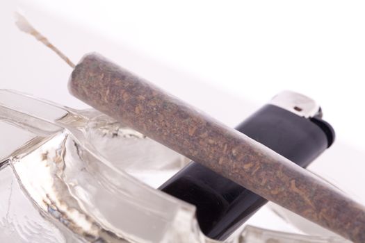 Close up of marijuana joint made with translucent rolling papers, plastic baggy of dried marijuana, black lighter and pipe on white background