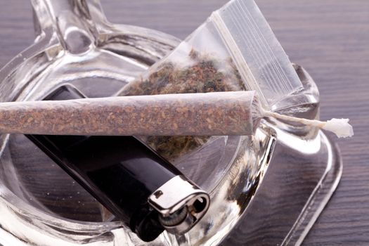 Close up of marijuana joint made with translucent rolling papers, plastic baggy of dried marijuana, black lighter and pipe on white background