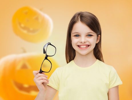 education, holidays, childhood, vision and people concept - smiling little girl in glasses over halloween pumpkins background