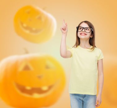 gesture, idea, education, holidays and people concept - smiling little girl in glasses pointing finger over halloween pumpkins background