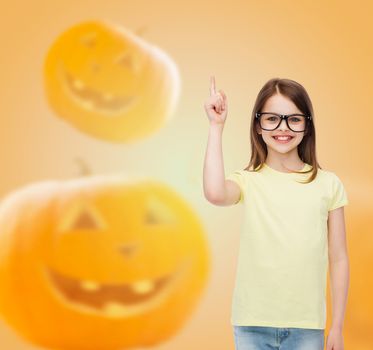 gesture, idea, education, holidays and people concept - smiling little girl in glasses pointing finger over halloween pumpkins background