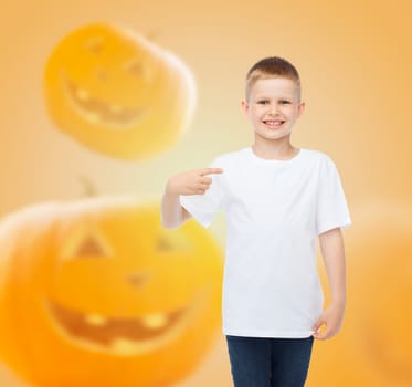 holidays, childhood, happiness, gesture and people concept - smiling little boy in white blank shirt pointing finger at himself over halloween pumpkins background