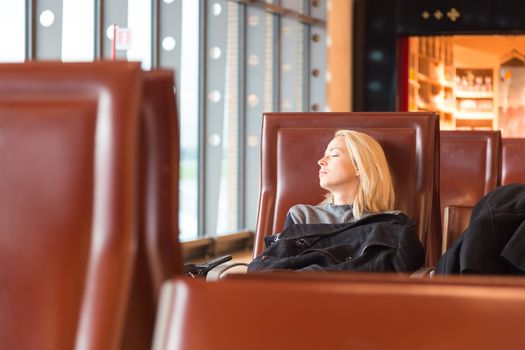 Cold and tired female traveler covered with jacket waiting for departure, resting on the gates bench with all her luggage by her side.  Tireing business travel.