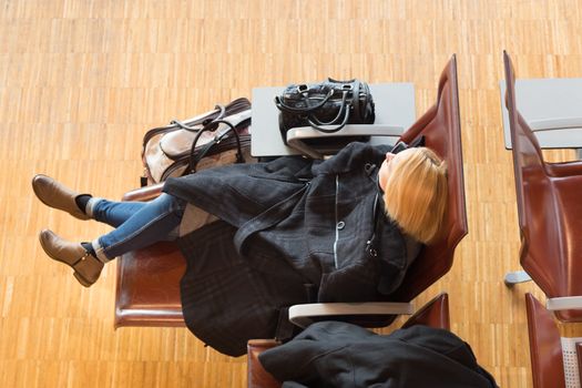 Cold and tired female traveler covered with jacket waiting for departure, resting on the gates bench with all her luggage by her side.  Tireing business travel.