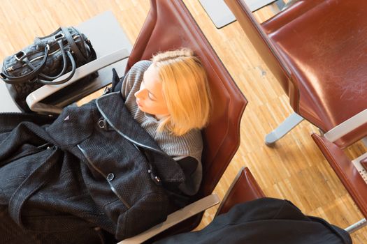 Cold and tired female traveler covered with jacket waiting for departure, resting on the gates bench with all her luggage by her side.  Tireing business travel.