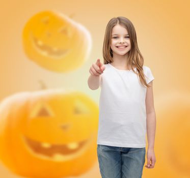 holidays, childhood, happiness, gesture and people concept - smiling little girl in white blank shirt pointing finger at you over halloween pumpkins background