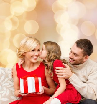 christmas, holidays, family and people concept - happy mother, father and little girl with gift box kissing over beige lights background