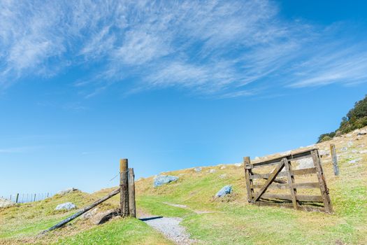 Open old wooden farm gate on hillside under blue sky with light wispy clouds