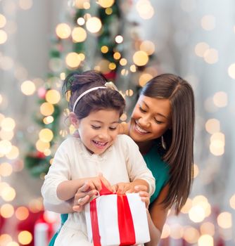 holidays, celebration, family and people concept - happy mother and little girl with gift box over living room and christmas tree background