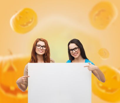 advertising, education, holidays and people concept - smiling teenage girls in glasses holding white board and pointing fingers over halloween pumpkins background