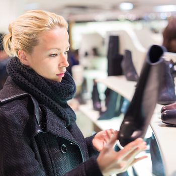 Woman shopping shoes. Shopper looking at ancle high boots on display shelf in shoe store. Beautiful blonde caucasian female model. 