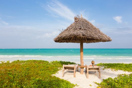 Women sunbathing on dack chair under wooden umbrella on stunning tropical beach. Turquoise blue lagoon of Paje beach, Zanzibar, Tanzania in the background.