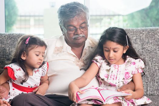 Grandparent and grandchildren reading story book. Happy Indian family at home. Asian grandfather and granddaughters indoor lifestyle.