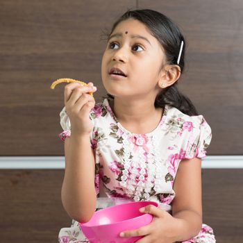 Cute Indian girl eating traditional snack murukku. Asian child enjoying food, living lifestyle at home.