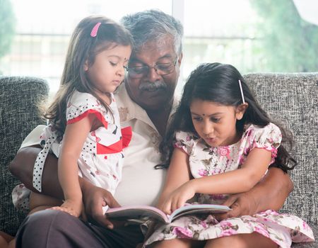 Asian grandfather and granddaughters reading story book. Happy Indian family at home. Grandparent and grandchildren indoor lifestyle.