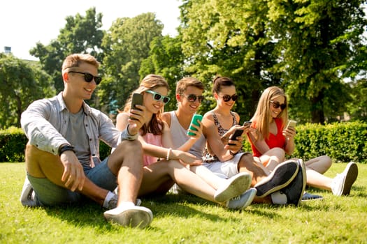 friendship, leisure, summer, technology and people concept - group of smiling friends with smartphones sitting on grass in park