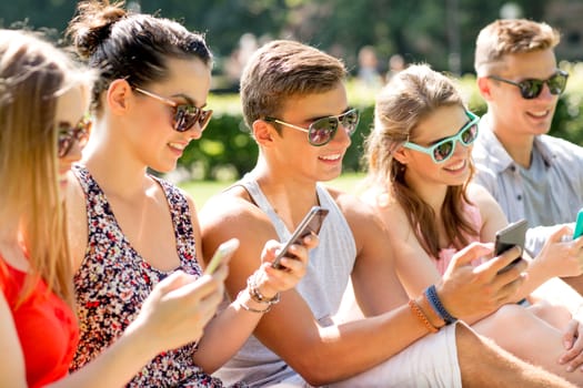 friendship, leisure, summer, technology and people concept - group of smiling friends with smartphones sitting on grass in park