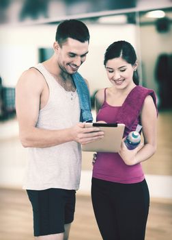 fitness, sport, training, gym, technology and lifestyle concept - two smiling people with tablet pc computer and water bottle in the gym