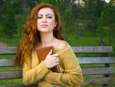 Ginger-haired woman sitting on a bench with book in the park
