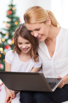 family, childhood, holidays, technology and people - smiling mother and little girl with laptop computer over living room and christmas tree background