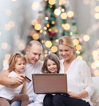 family, holidays, shopping, technology and people - happy family with laptop computer and credit card over christmas lights tree background