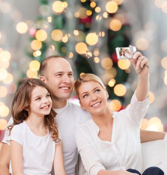 family, holidays, technology and people - smiling mother, father and little girl making selfie with camera over christmas tree lights background