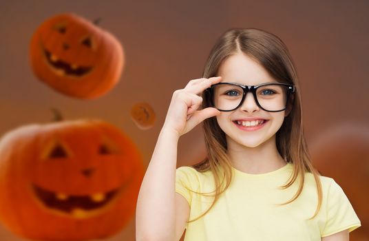 education, holidays, childhood, vision and people concept - smiling little girl in glasses over halloween pumpkins background