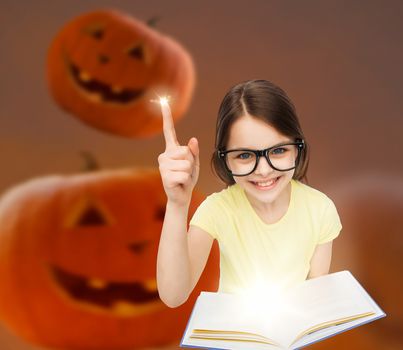 education, holidays, childhood, vision and people concept - smiling little girl in glasses with magic book over halloween pumpkins background