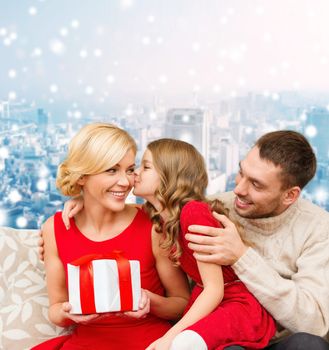 christmas, holidays, family and people concept - happy mother, father and little girl with gift box kissing over snowy city background