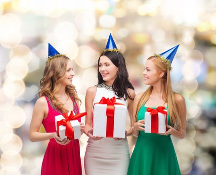 presents, holidays, people and celebration concept - smiling women in party caps with gift boxes over lights background