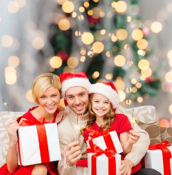 holidays, family and people concept - happy mother, father and little girl in santa helper hats with gift boxes and sparklers over living room and christmas tree background
